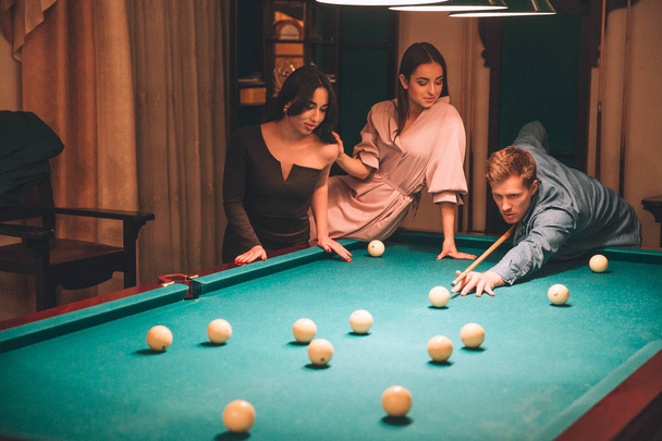 Concentrated young redhead man aiming into billiard ball. He look straight forward. Young woman in pink dress sit at billiard table. Asian brunette stand beside them. Women look at game. - Photo, image