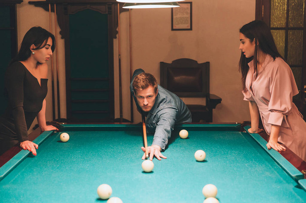 Young redhead player stand between two models. He aims into billiard ball. Young women stand and look at gamel. They lean to table. - Foto, immagini