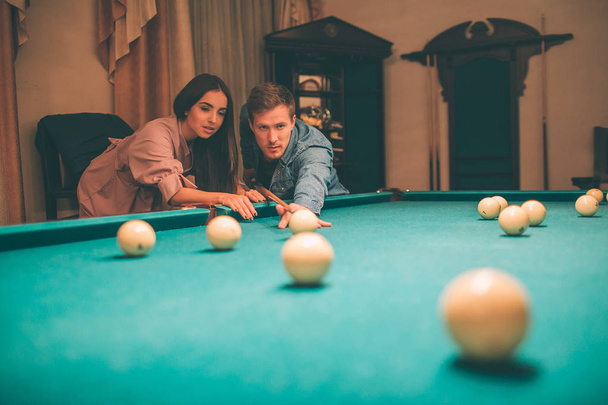 Young player stand in angled position and aiming to break ball. He looks concentrated. Brunette stand besides him and look at billiard balls. Guy hold cue in hands. THey are in playing room. - Photo, image