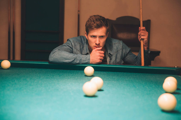 Serious and concentrated young man thinking. He look at billiard balls on bed of table. Guy hold billiard cue. He is alone in room. - Photo, image
