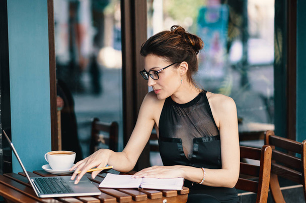 Femme et technologie. Femme d'affaires raffinée dans des lunettes, assise à table dans un café avec des fenêtres en arrière-plan prenant des notes sur un carnet. Sur la table est ordinateur portable, ordre du jour et une tasse de café
 - Photo, image