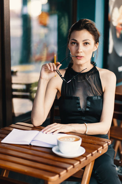 Elegante jonge vrouw nam haar bril terwijl wordt gekeken naar de camera glimlachen, ze hebben een pauze van haar agenda zittend op het terras van een café met een grote kop koffie met windows op achtergrond. - Foto, afbeelding