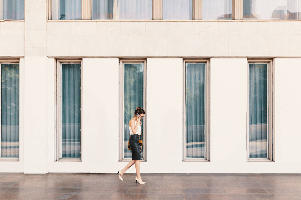 Exquisite business woman in leather skirt and silk blouse talking on the phone while walking near a building with big windows. Business, communication, technology and people concept. - Foto, Imagen