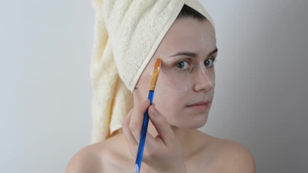 Attractive young woman wrapped with bath towels, applying Cream on her Face with a brush After a Shower. Skin care and beauty concept. Applying facial mask at woman face at home. - Felvétel, videó