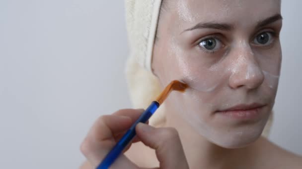 Attractive young woman wrapped with bath towels, applying Cream on her Face with a brush After a Shower. Skin care and beauty concept. Applying facial mask at woman face at home. - Filmagem, Vídeo