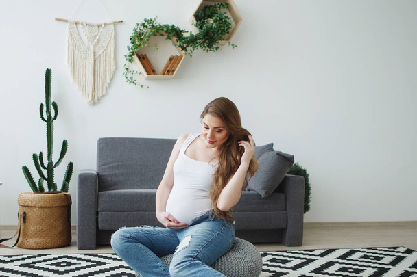 Красива вагітна жінка з довгим волоссям у білому жилеті сидить на підлозі біля сірого дивана на картатому килимі. Концепція щасливого материнства, здоровий спосіб життя
. - Фото, зображення