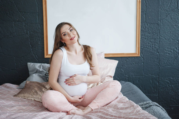 Beautiful pregnant woman with long hair in a white T-shirt and pink pajamas is sitting on the bed and stroking her tummy. Concept of happy motherhood, healthy lifestyle. - Fotó, kép