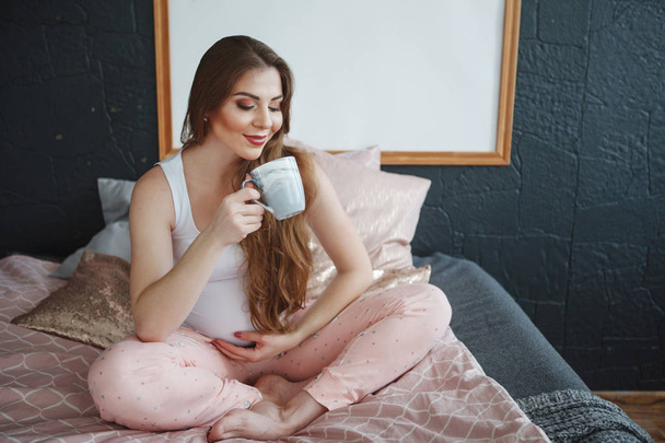 Beautiful pregnant woman with long hair in a white T-shirt and pink pajamas sitting on the bed, holding a cup of tea. Concept of happy motherhood, healthy lifestyle. - Фото, зображення