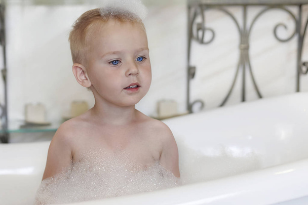 Cute caucasian baby boy with bright blue eyes and fair hair in the bathtube, with foam bubbles o the head and around. Indoors, copy space. - Fotó, kép