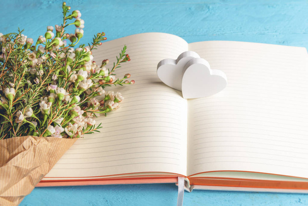 Pequeno buquê de flores brancas em um caderno vazio e dois corações de madeira, em fundo azul. Presente do dia dos namorados. Contexto de momentos felizes. Aniversário
. - Foto, Imagem