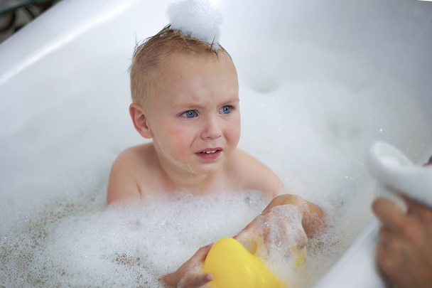 Cute caucasian baby boy with blue eyes in the bathtube cries because of foam bubbles on his face and head; yellow toy duck in his hands. Mother' hands with towel. Indoors, close up portrait, copy space. - Foto, Bild