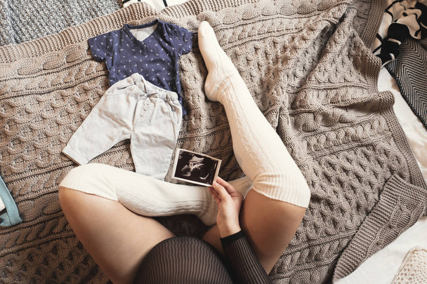 Spodziewając się kobieta w ciąży dotykając jej brzuch, patrząc na USG, siedząc na wygodnym łóżkiem, koce, poduszki, ubrania dziecko, smartphone, noszenie długie białe skarpetki. Widok z góry przycięte zdjęcie. - Zdjęcie, obraz