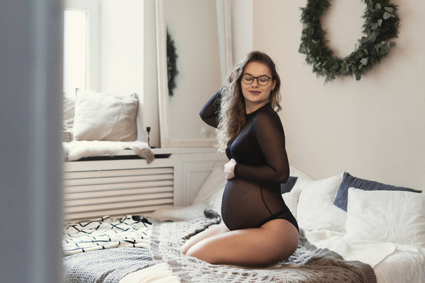 Beautiful pregnant woman, wearing transparent black body lingerie enjoying her baby moves while touching her belly sitting on bed at home. Peoples emotions, waiting a new life. - Photo, Image