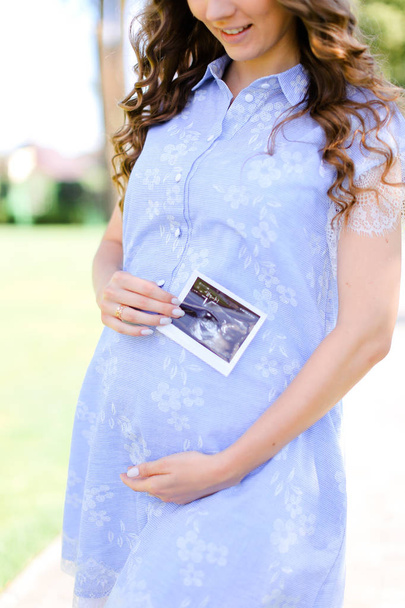 Happy young pregnant woman wearing blue dress holding belly with ultrasound. - Photo, Image