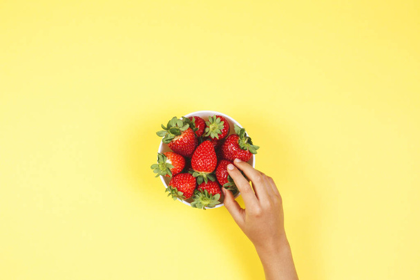 Kid hand taking strawberry from the bowl on yellow background - Zdjęcie, obraz