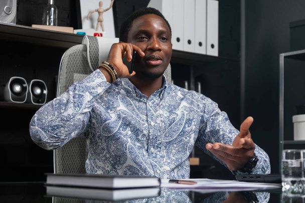 молодой черный бизнесмен разговаривает по мобильному телефону сидя за компьютером стол в офисе
 - Фото, изображение