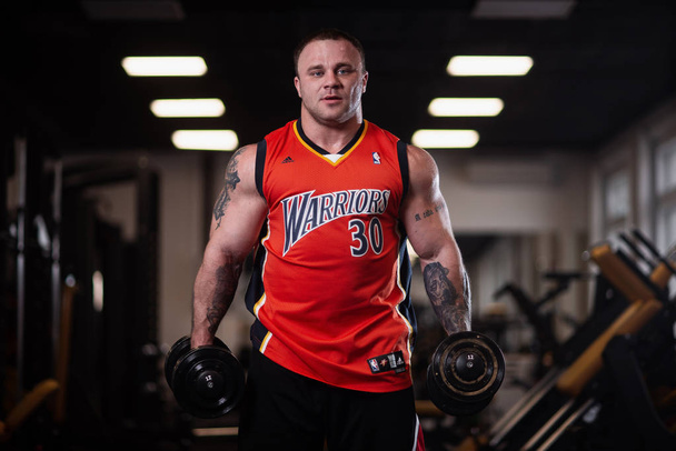 RUSSIA, SAMARA - DECEMBER 18, 2018: Big athletic man trains in the gym. Training biceps with a dumbbells - 写真・画像