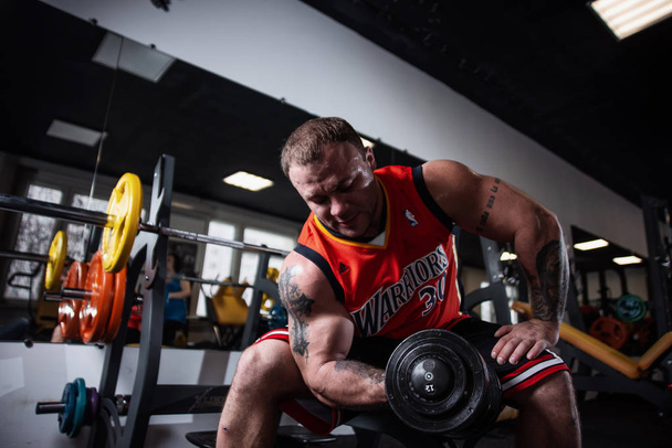 Rusland, Samara - 18 December 2018: Een grote atletische man training in de sportschool. Opleiding van biceps met halters, zittend op een bankje - Foto, afbeelding