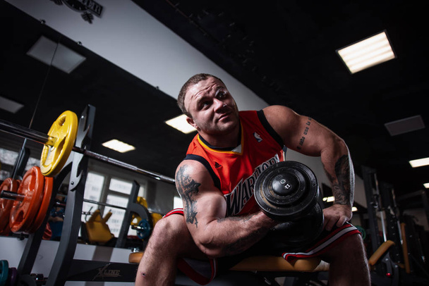 RUSSIA, SAMARA - DECEMBER 18, 2018: A large athletic man training in the gym. Training of biceps with dumbbells, sitting on a bench - 写真・画像