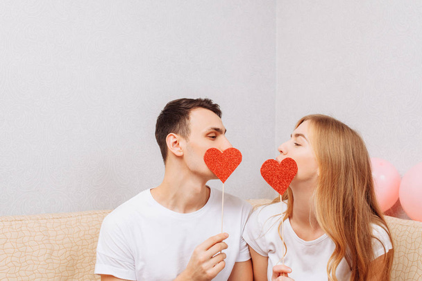 Beautiful couple in love, man and woman in white T-shirts, holding paper hearts, sitting on the sofa - Fotoğraf, Görsel