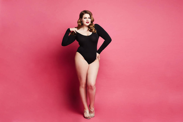 Plus size sexy model girl, fashionable blonde in black bodysuit, with bright makeup and with stylish hairstyle, smiling and posing at pink background in studio - Photo, Image