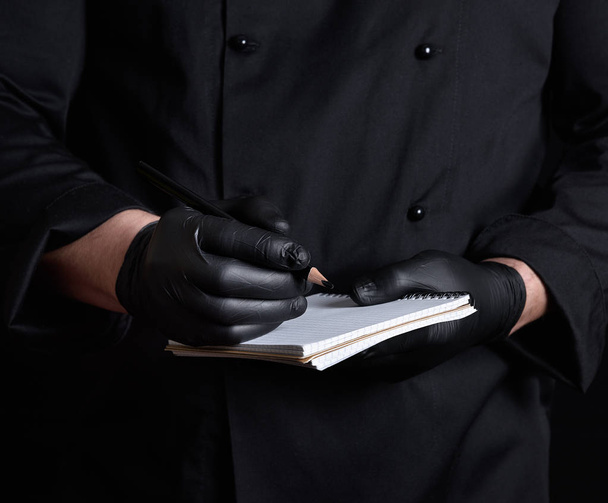 chef in black uniform and latex gloves holding a blank notebook and a black wooden pencil, close up - Foto, Imagem