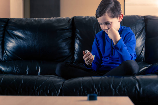 Ragazzo di 8 anni che indossa abbigliamento sportivo guardando il suo inalatore d'asma seduto sul divano. Concetti: salute, malattia, doping
 - Foto, immagini