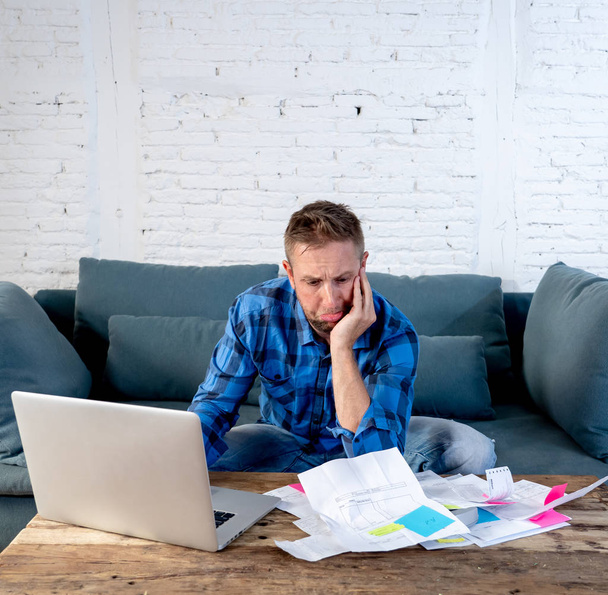 Worried and stressed man calculating bills tax expenses and counting home and business finances sitting on the couch at home in paying off debts and domestic bills and financial problems concept. - Foto, imagen