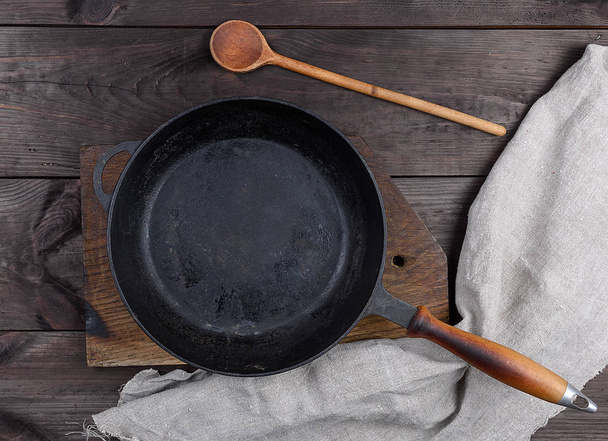 пустая черная круглая сковорода с деревянной ручкой и деревянной ложкой на коричневом столе, вид сверху
 - Фото, изображение