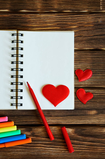 Blank notepad with felt-tip pens and red hearts on wooden background. Preparation for congratulations on Valentine's Day. Template. Vertical image.                              - Fotografie, Obrázek