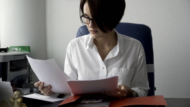 Крупным планом для молодой женщины в белой рубашке, смотрящей на ноутбук, отвечающей на звонки и улыбающейся на белом фоне офисной стены. Деловая женщина, работающая перед экраном компьютера на своем рабочем месте
. - Фото, изображение