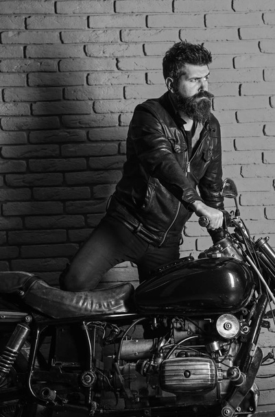 Hipster, brutal biker on serious face in leather jacket gets on motorcycle. Masculine passion concept. Man with beard, biker in leather jacket near motor bike in garage, brick wall background - Photo, image