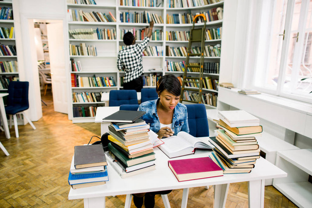 Africano americano jovem leitura livro sentado à mesa no fundo da estante. Concentrado escuro pecado estudante passar o tempo de lazer na biblioteca
 - Foto, Imagem