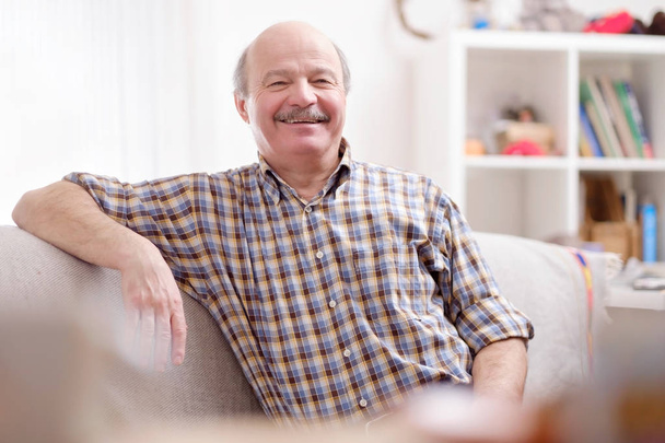 Bonito homem hispânico maduro sentado no sofá em sua sala de estar sorrindo. Emoção facial positiva na velhice
 - Foto, Imagem