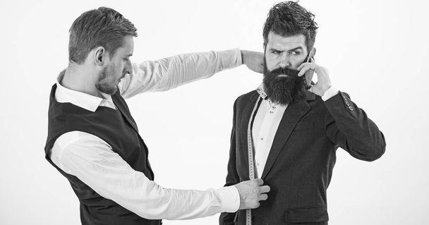 Man with beard holds measuring tape. Business clothing concept. Tailor measures chest for sewing, white background. Businessman, client on busy face speak on phone, smartphone while tailor working. - Foto, Imagem