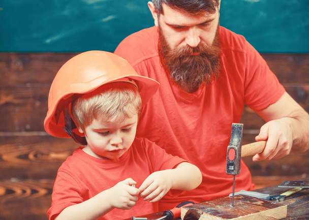 Father, parent with beard teaching little son to use hobnails and hammer. Boy, child busy in protective helmet learning to hammering hobnails with dad. Masculine duties concept - Photo, image
