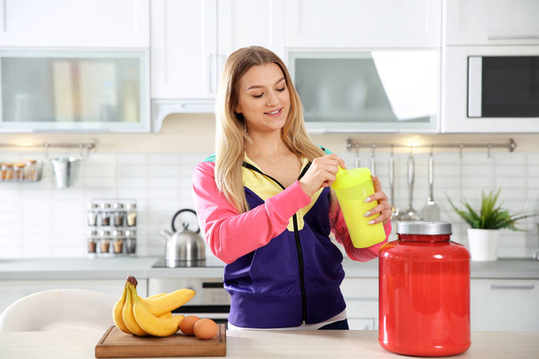 Young woman holding bottle of protein shake near table with ingredients in kitchen - Фото, зображення