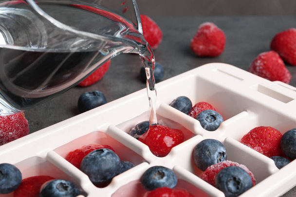 Pouring water into ice cube tray with berries on table, closeup - 写真・画像
