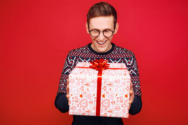 Happy guy in glasses with a gift box in his hands, looks at her, on a red background - Foto, Imagen