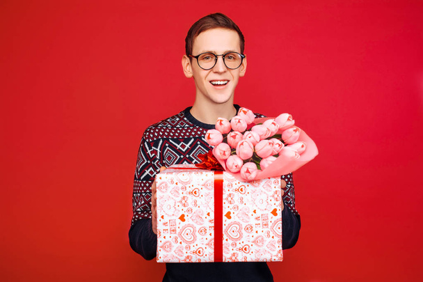 man in glasses, man with a gift and a bouquet of flowers in his hands, on a red background - Foto, afbeelding