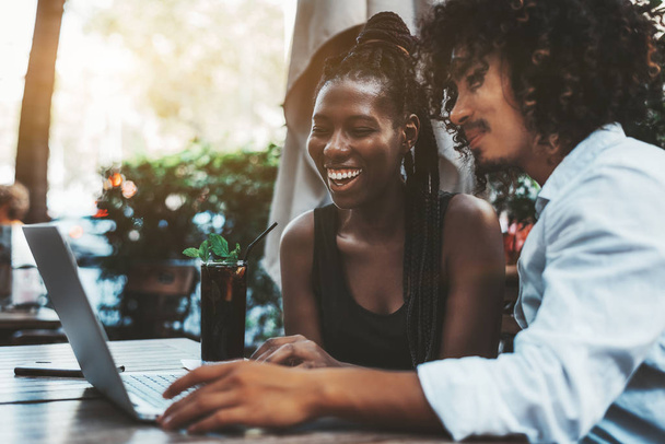 A dazzling laughing black girl with a braided hair is looking on the screen of the laptop where her Asian bearded curly male boyfriend is showing her photos from the friends party, glass of drink near - Zdjęcie, obraz