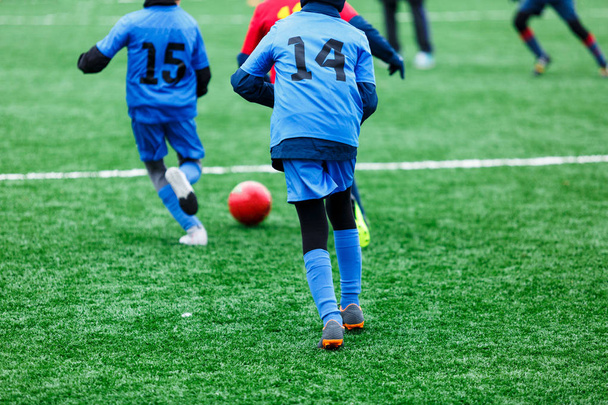 Мальчики в красно-синей спортивной одежде играют в футбол на зеленом травяном поле. Молодежный футбол. Детские спортивные соревнования, детские игры на свежем воздухе, зимние мероприятия, тренировки
 - Фото, изображение