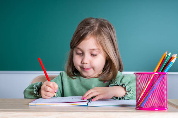 kleines hübsches Mädchen sitzt im Klassenzimmer am grauen Schreibtisch und zeichnet. - Foto, Bild