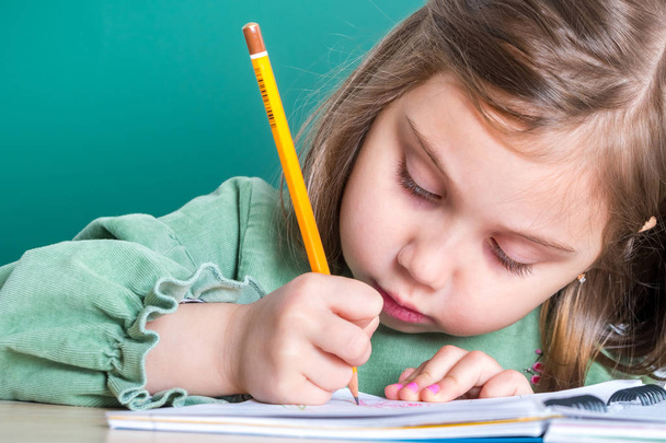 kleines hübsches Mädchen sitzt im Klassenzimmer am grauen Schreibtisch und zeichnet. - Foto, Bild