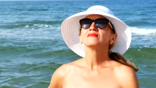    4 k. Pretty volwassen vrouw in witte hoed en bril, staande in de buurt van de oceaan golven.  - Video