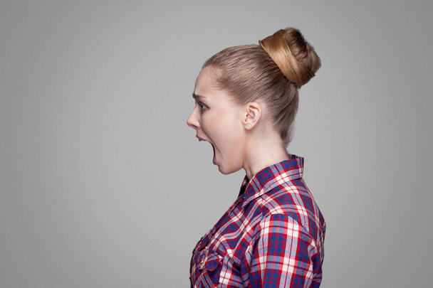 side view of scared blonde woman in checkered shirt with collected bun hairstyle screaming on gray background - Foto, Imagem