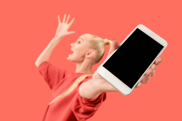 Portrait of a confident casual girl showing blank screen mobile phone isolated over coral background - 写真・画像