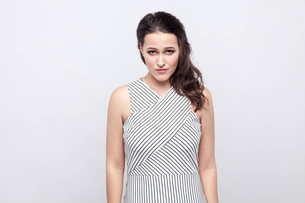 Portrait of sad beautiful young brunette woman with makeup and striped dress standing at camera with bad feelings and depressed sadness eyes on grey background. - Photo, Image
