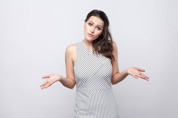 Confused young brunette woman with makeup and striped dress standing and looking at camera with raised arms on grey background - Foto, Bild