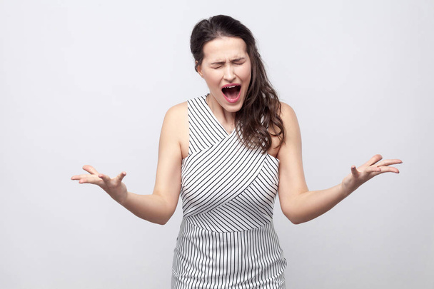 Portrait of angry young brunette woman with striped dress standing with crossed arms and screaming on grey background - Foto, imagen
